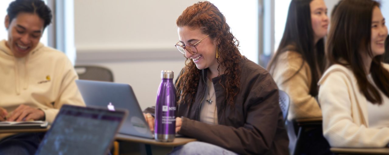 An NYU SPS student looks down at her computer and smiles while participating in a class discussion.