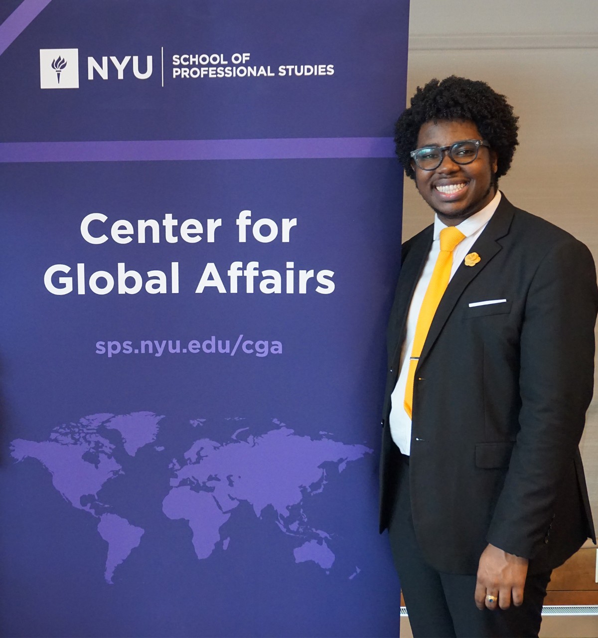 A smiling person in a suit with a bright yellow tie stands next to a purple NYU School of Professional Studies banner for the Center for Global Affairs.