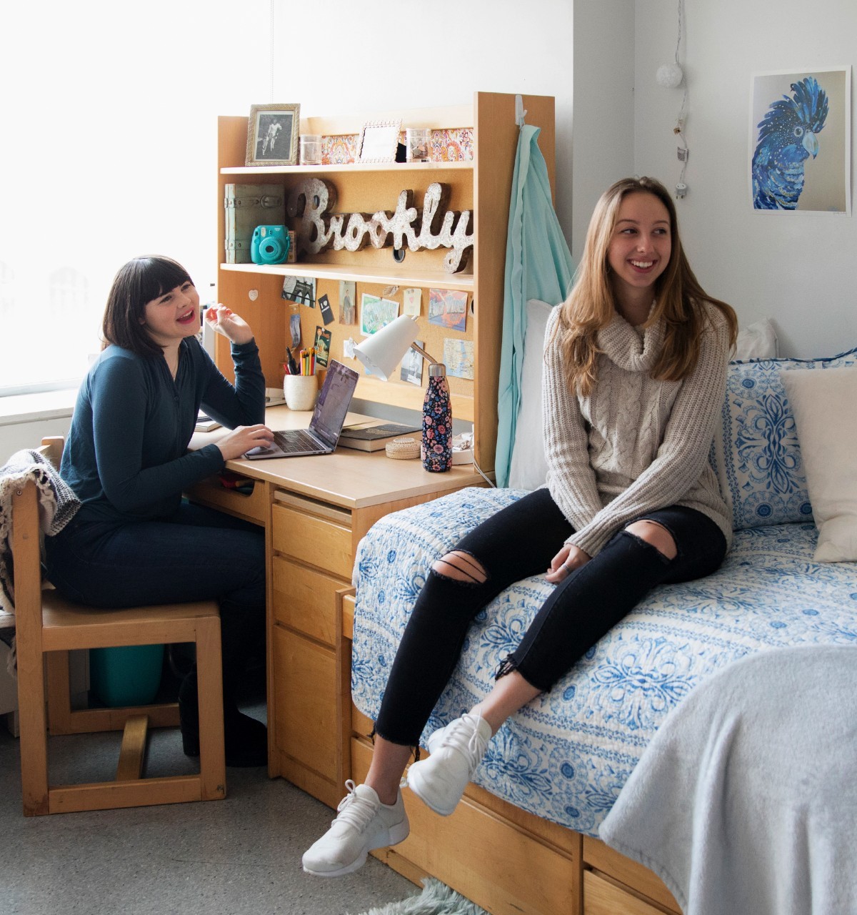 Two students smiling in a dorm room, one sitting on a bed and the other at a desk with a laptop