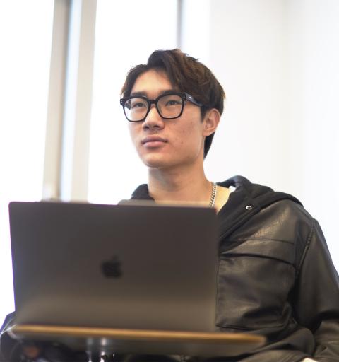 An NYU SPS student sits at a desk behind a laptop during an online language proficiency testing session