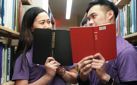 Two students reading a book together in a library aisle
