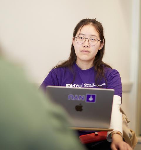 Young woman in a purple NYU shirt attentively looks at a laptop in a classroom setting.