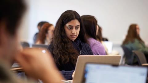 A student with long dark hair focuses on her laptop during class, surrounded by other students in a bright classroom setting.