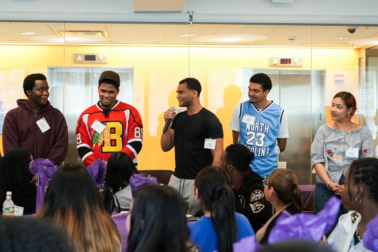 A group of five young adults stands in front of an attentive audience in a bright room. One person holds a microphone, suggesting a presentation or speech. They are all smiling, dressed casually in sports jerseys and casual wear.