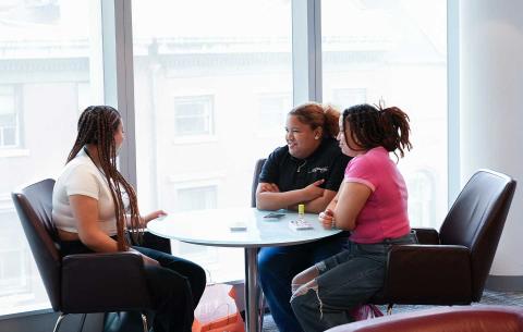 Three people sitting around a table having a conversation in a well-lit room with large windows.