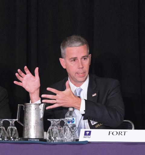 A speaker at an NYU SPS conference gestures with his hands while making his point on stage.