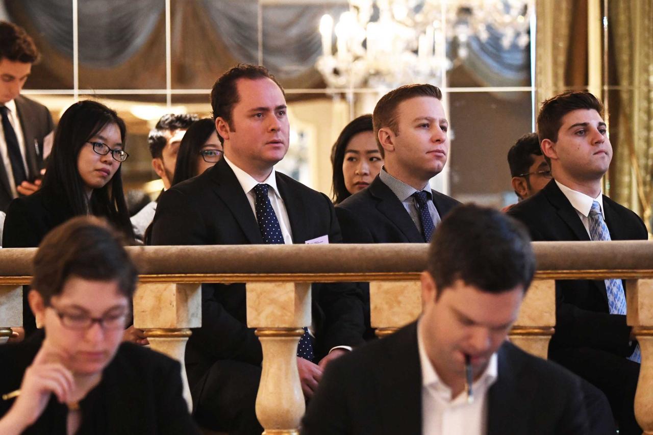 A group of people in formal attire sit attentively in an elegant conference room with chandeliers and mirrors.