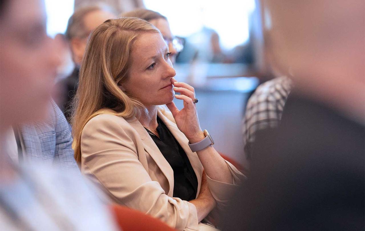 A woman with long blonde hair, wearing a beige blazer, attentively listens in a conference.