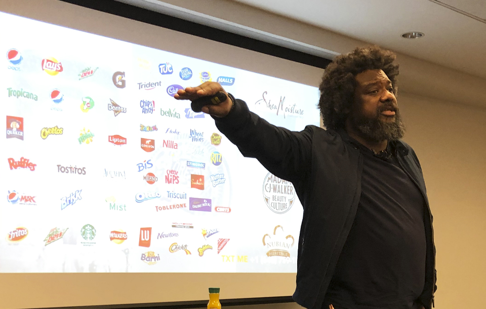 An NYU SPS instructor provides remarks in front of a projected slide containing dozens of corporate logos.