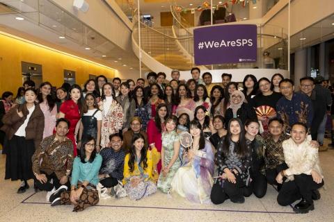 A diverse group of NYU SPS students in cultural attire gathers at an event, celebrating community and inclusion under a "#WeAreSPS" sign