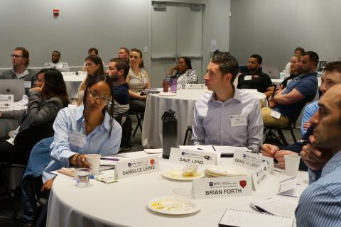 Attendees seated at round tables during a conference, focusing on a presentation.