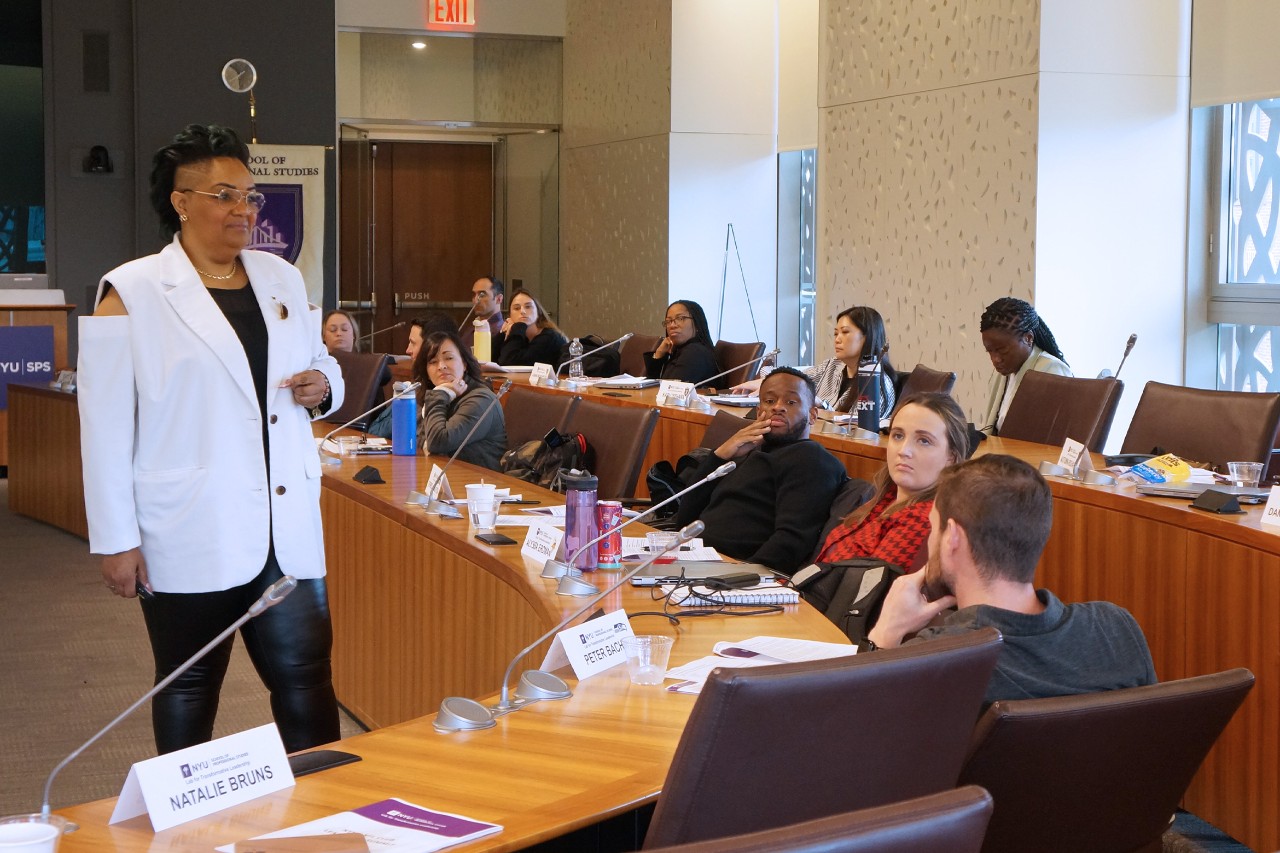 A speaker in a white blazer addresses attendees in a conference room.