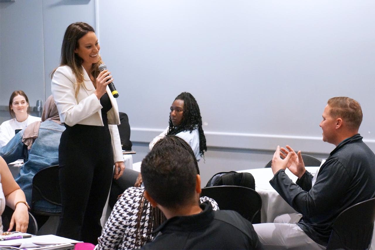 A woman with a microphone speaks to a seated man in a conference room. Other attendees are seated at tables.