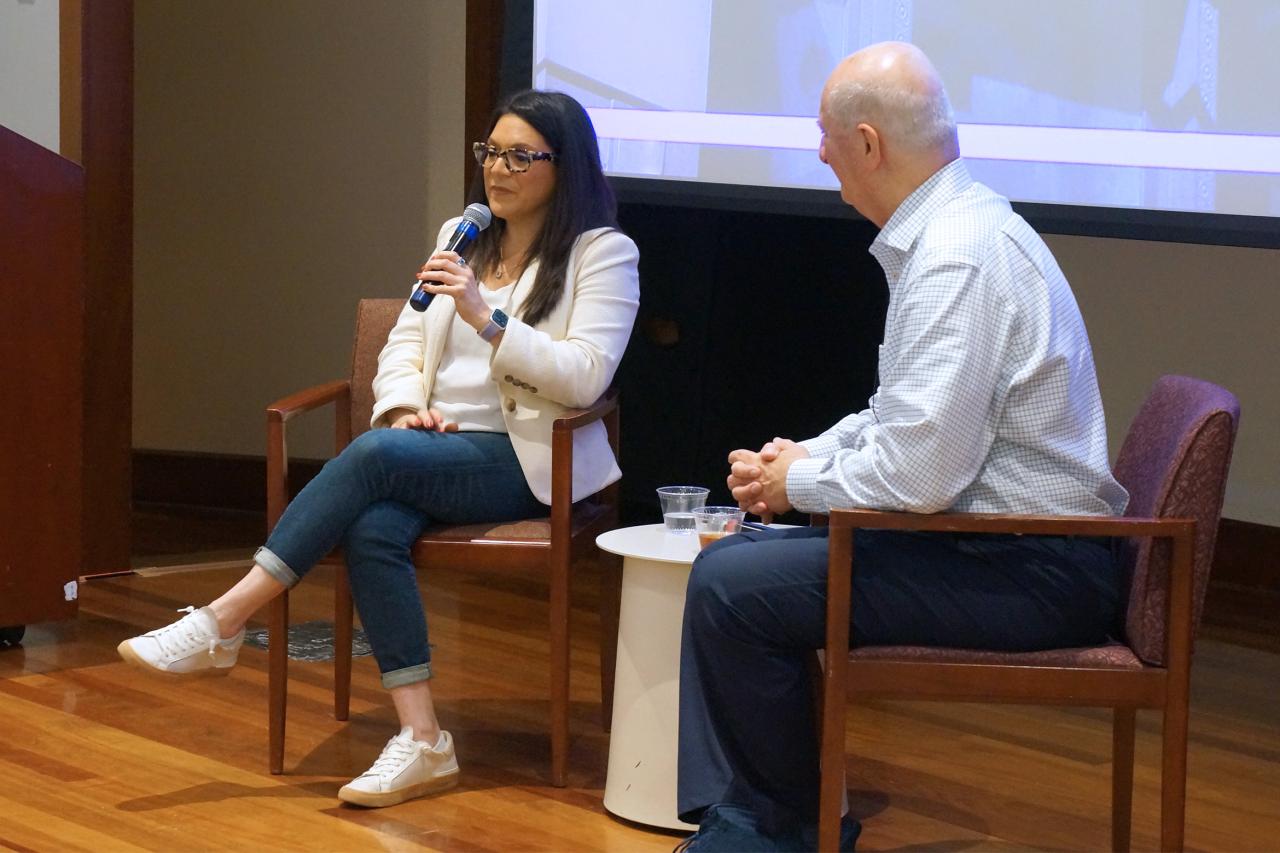 Two people seated in a conference room during a panel discussion, one holding a microphone, with a podium and projection screen in the background.