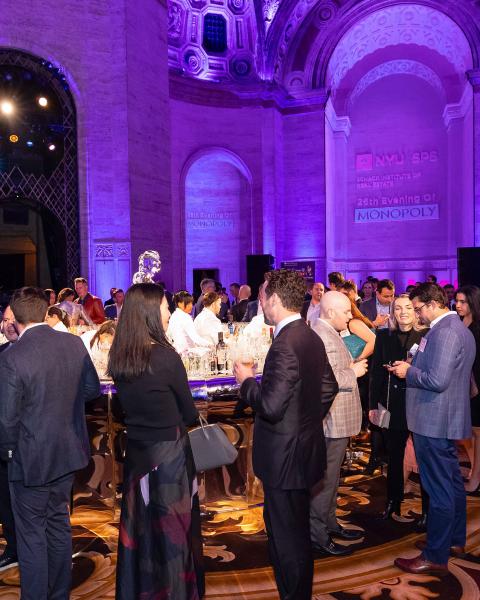 People socializing at NYU SPS Schack Institute of Real Estate 26th Evening of Monopoly in a grand room with purple lighting.