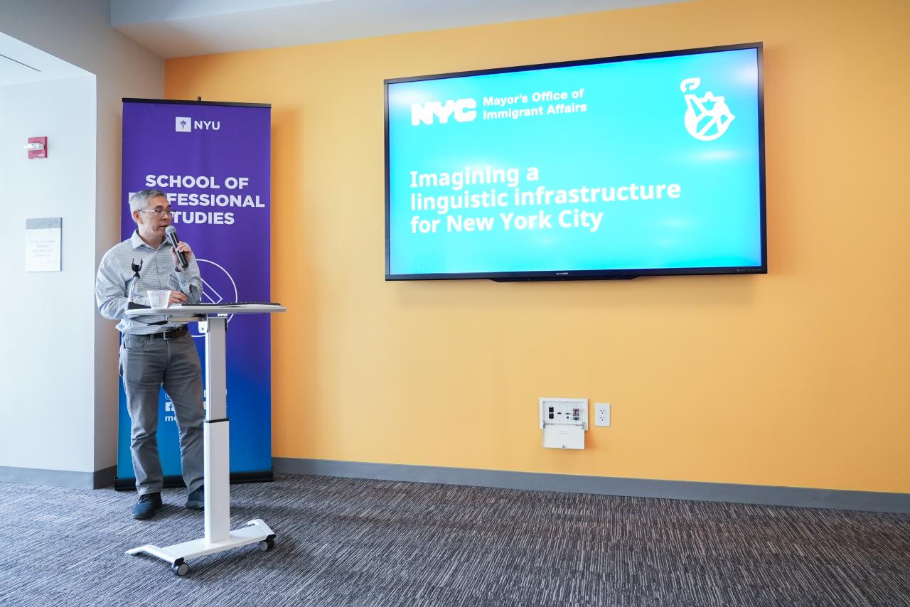 Man in slacks and long sleeve plaid tee standing at a podium in front of a TV powerpoint that reads &quot;Imagining a linguistic infrastructure for New York City&quot;