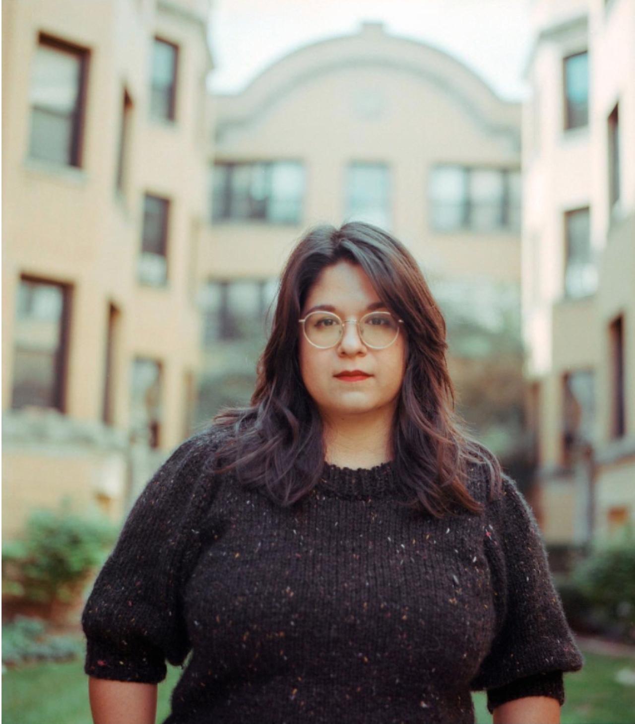 Front profile of Alejandra Oliva as they stand outside in front of a blurred beige building. They are female presenting with shoulder length chocolate brown hair, brown eyes, and gold, round classes wearing a blackish sweater