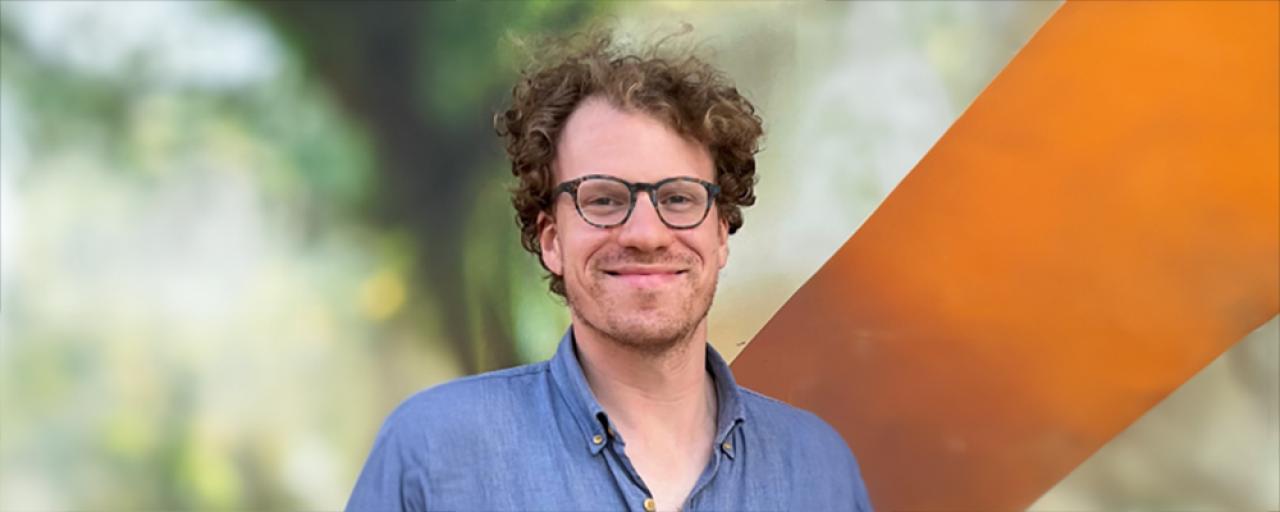 Picture of Justin Sergi (light brown curly hair, glasses) as they look at the photographer with the background blurred. This picture is placed on a light purple, textured background