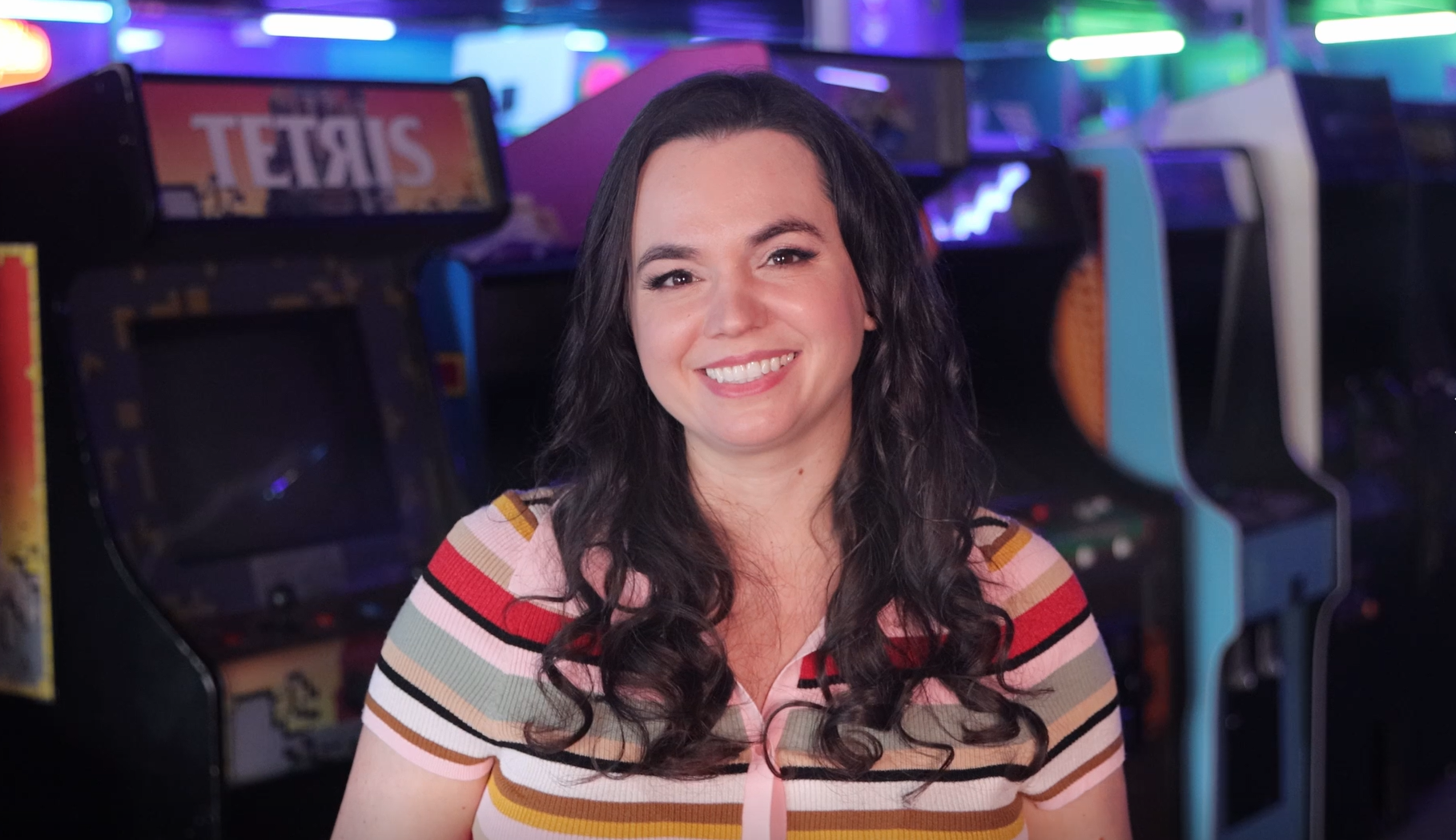 Image of Marina Ilari. Marina is woman presenting with long brown hair and brown eyes. They are wearing a colorful striped shirt and standing in front a row of video game machines.