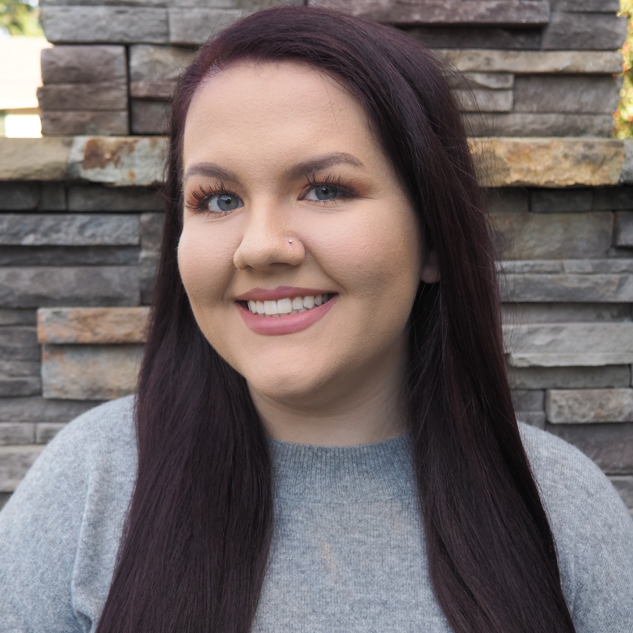 Image of Olivia Langenberg. Olivia presents as a woman and has long brown hair and blue eyes. They are wearing a gray shirt and are standing against a wood panel wall. 