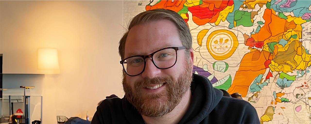 Front profile of Patrick Gray in front of colorful wall. Man with black glasses, blue eyes, and red-gray hair and beard smiling at camera.