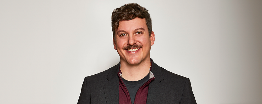 Image of Simon Mullin as they stand and face the camera. Simon is male presenting, curly brown hair and mustache, brown eyes.
