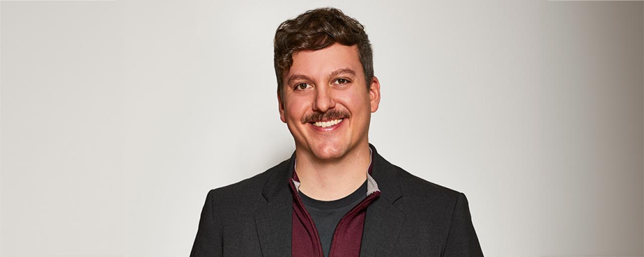 Image of Simon Mullin as they stand and face the camera. Simon is male presenting, curly brown hair and mustache, brown eyes.