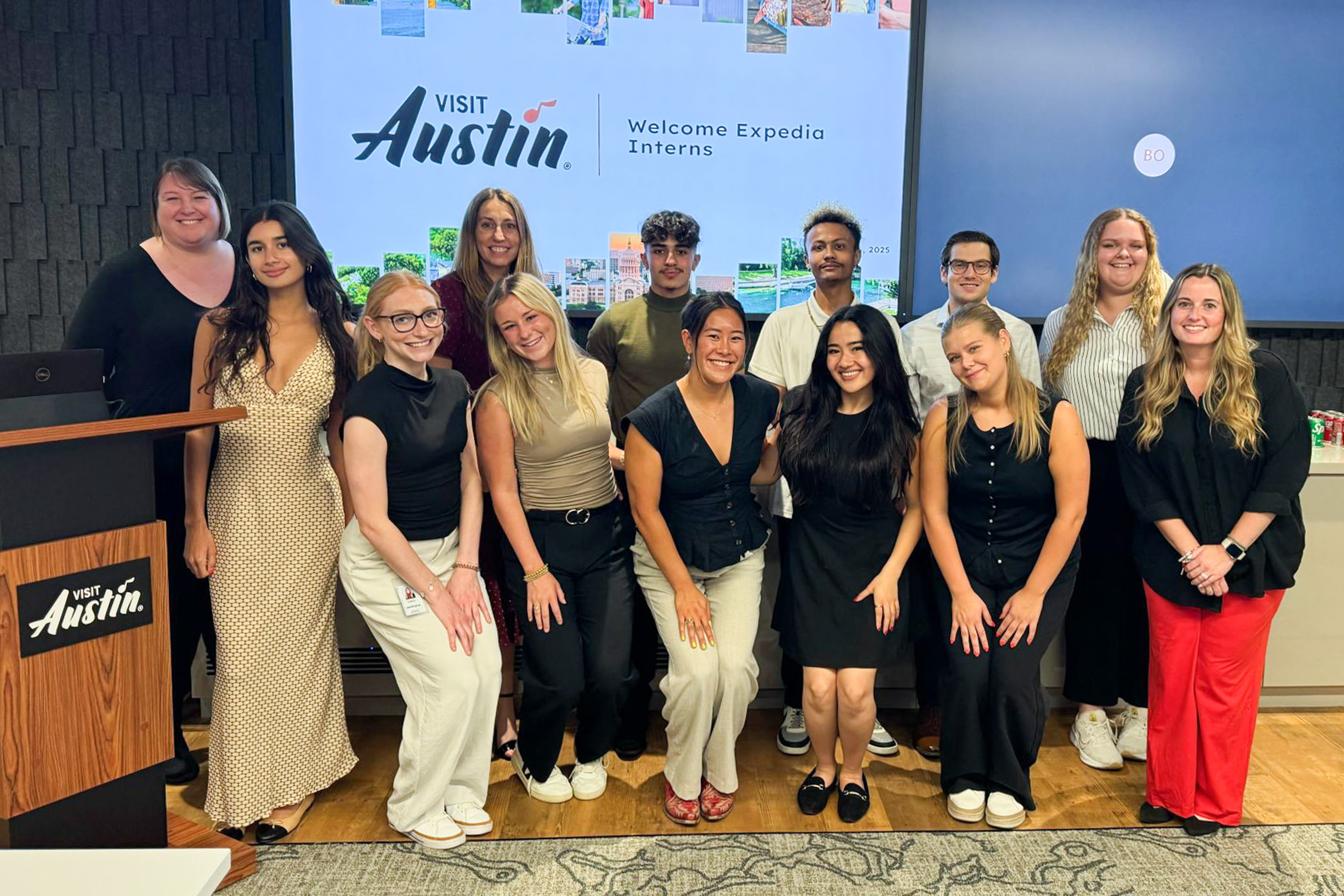 A group of thirteen people posing in front of a screen displaying a welcome message for Expedia Interns.