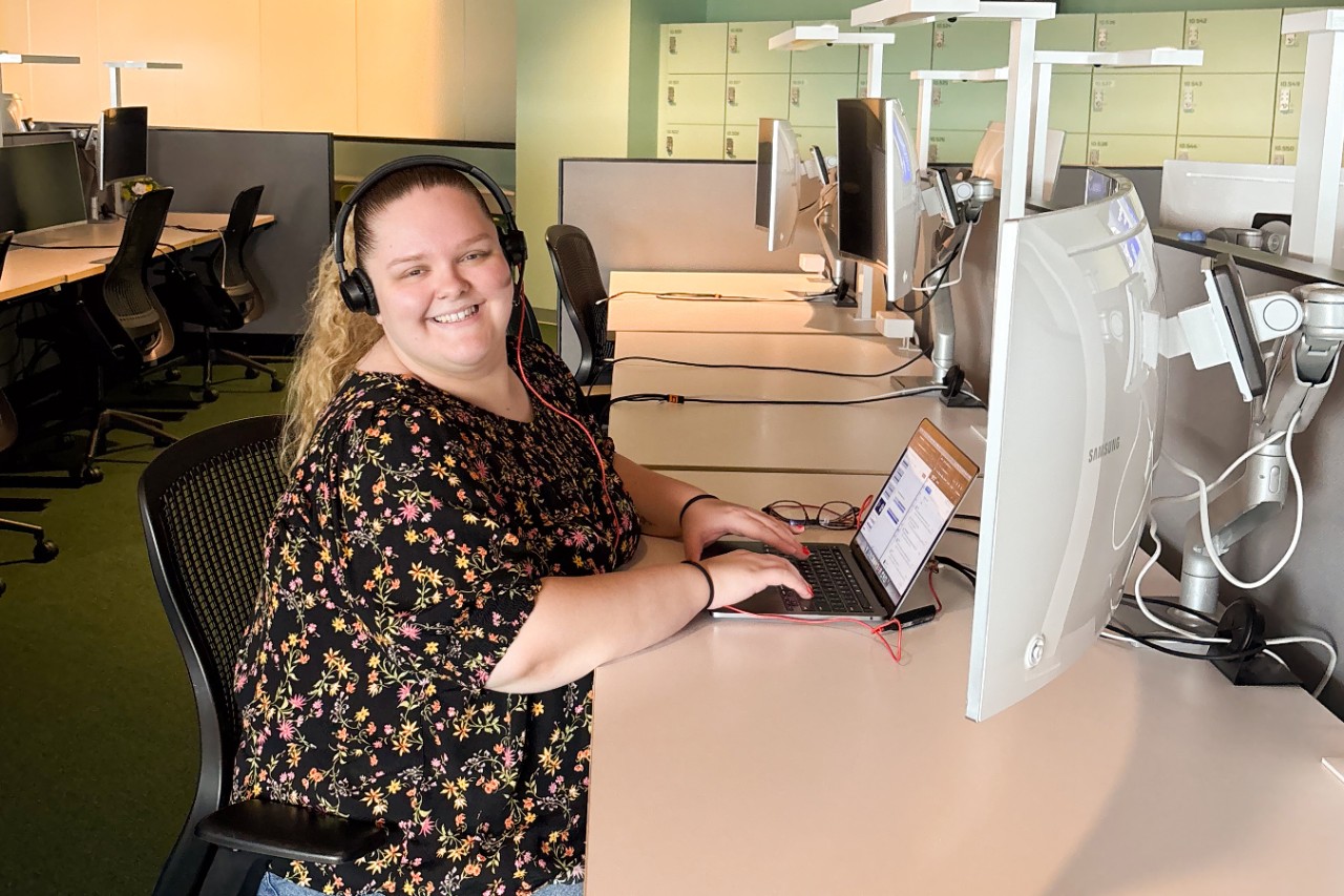 A person with headphones working on a laptop at a desk in a modern office with exposed ceilings and light green walls.