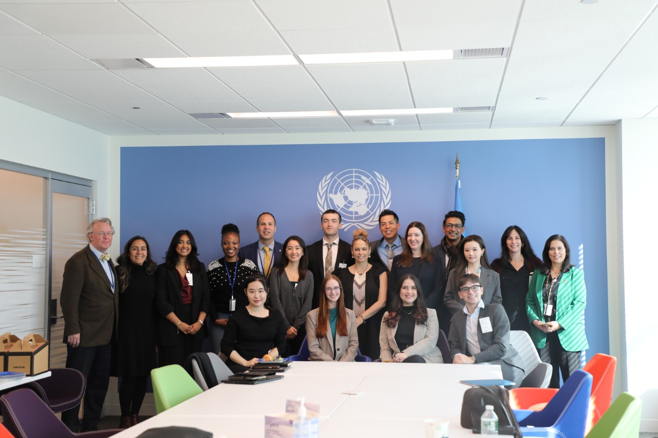 Group photo of students and faculty standing and seated together in a United Nations conference room, posed in front of the UN emblem.