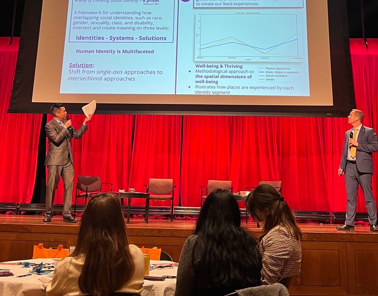 Two presenters standing on stage speaking in front of a projected slide during the Cracking the Connection Code event.