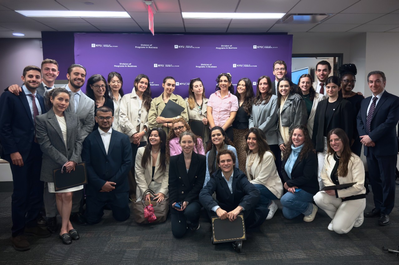 Group photo of diverse adults in business attire smiling in a conference room at NYU. A purple backdrop with text is in the background.