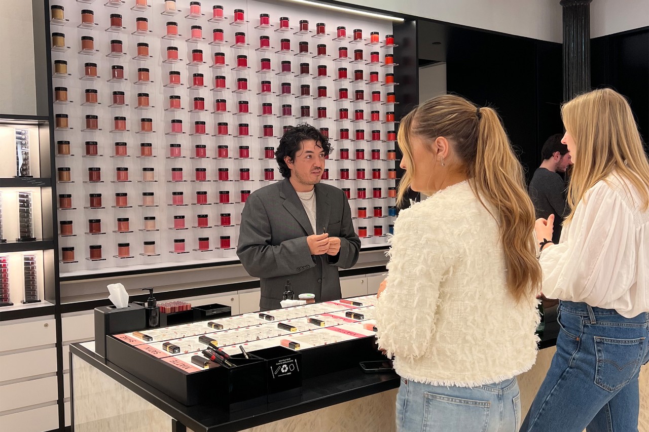 Two women with long hair, wearing jeans and light tops, talk to a salesperson at a makeup counter.