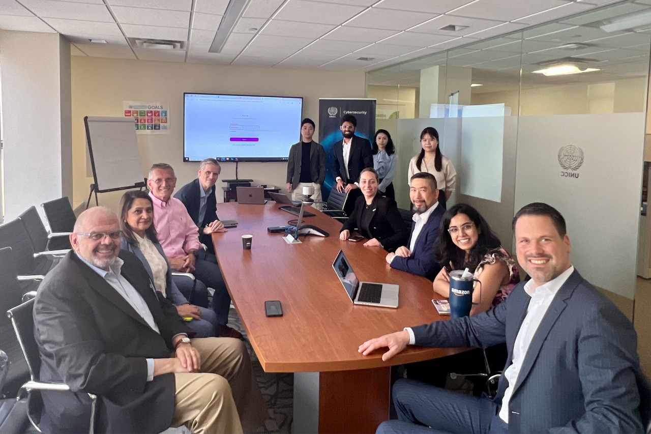 A group of twelve people in business attire gather around a conference table in a modern meeting room. Laptops and a presentation screen are visible.