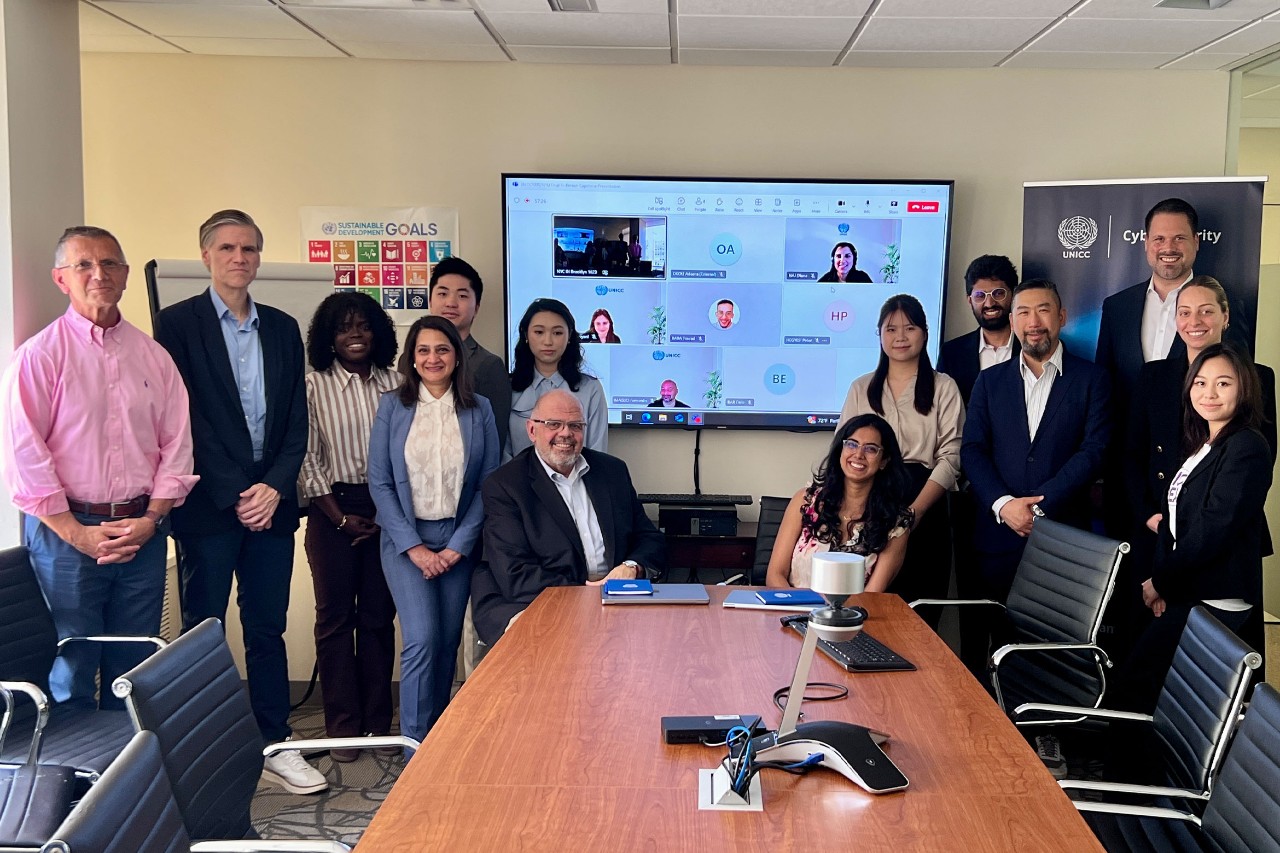 A diverse group of 14 people, dressed in business attire, pose around a conference table. A video conference call is displayed on the large screen behind them.