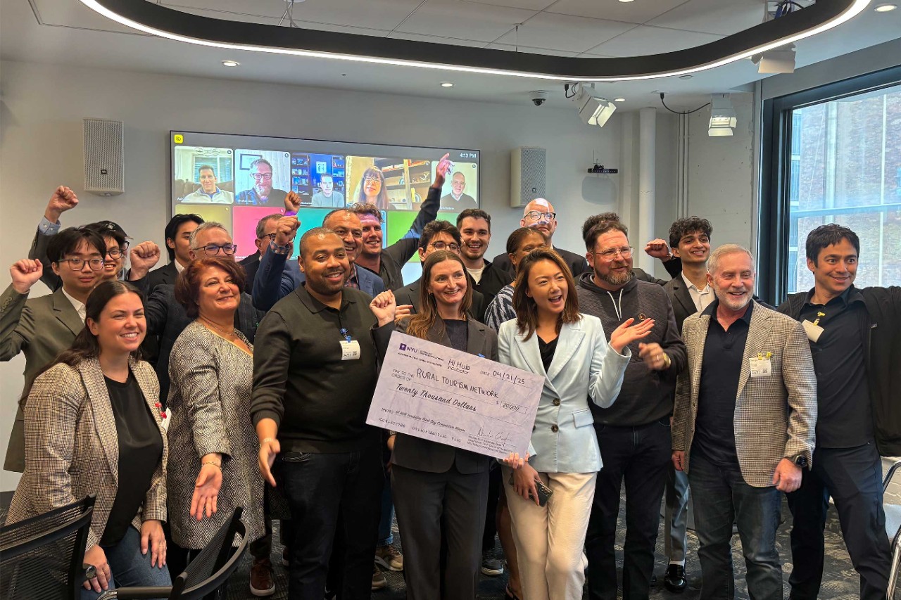 A diverse group of people cheerfully poses with a large check reading "Rural Tourism Network, Twenty Thousand Dollars." Virtual participants are visible on a screen behind them.