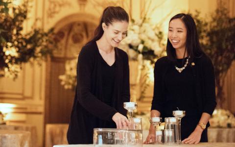 Two women dressed in black, smiling as they arrange glass candle holders on a table in an elegant, warmly lit event space decorated with flowers.