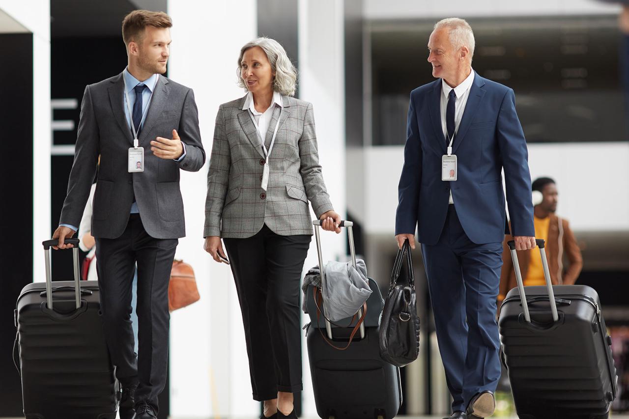 Confident team of forum participants in formalwear carrying wheeled suitcases and crossing airport together of guide