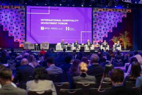 A conference panel on stage with five speakers seated, discussing under a bright purple backdrop displaying "International Hospitality Investment Forum" text. 