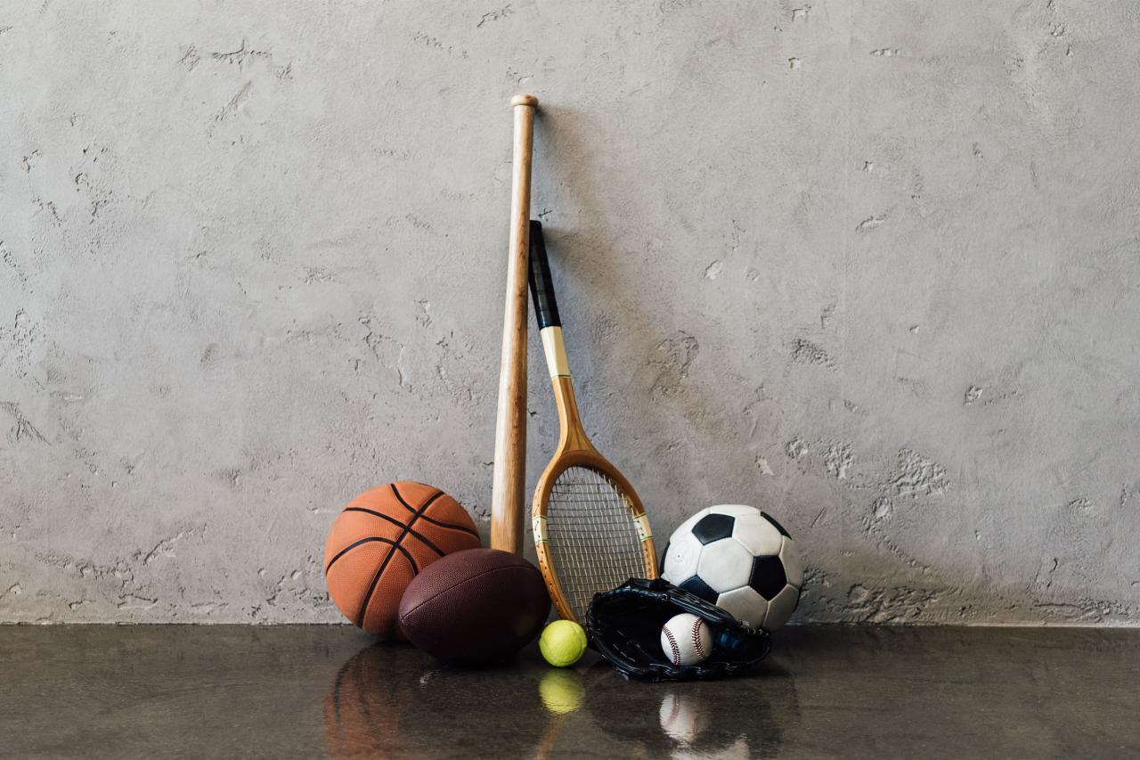 Basketball, tennis ball, football, soccerball, raquet, baseball bat, and glove against a grey wall.