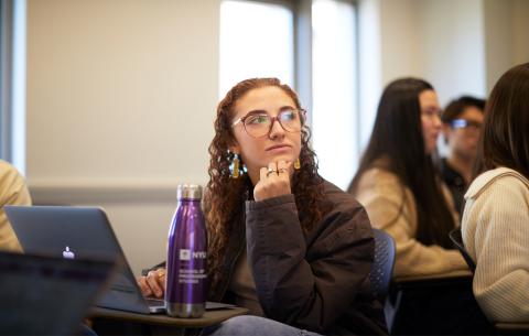 A female NYU SPS student sits at a desk behind her laptop and purple water bottle while listening to a lecture