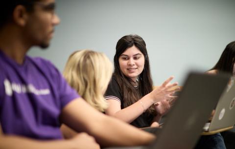Three master's degree students listen to a lecture while sitting at their desks behind their laptops and two of them speak to one another