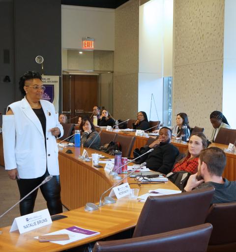 Interior of a conference room with a presenter in a white blazer addressing attendees seated at a curved table.