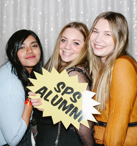 Three NYU SPS Class of 2024 alumni pose for a photo in a photobooth holding an "SPS Alumni" banner