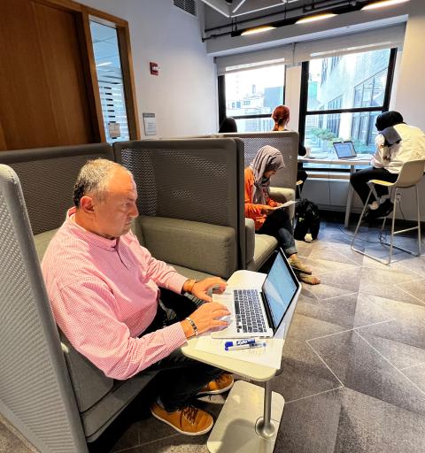 A man in a pink shirt works on a laptop in a modern, well-lit office space. People in the background are focused on their laptops, creating a studious atmosphere.