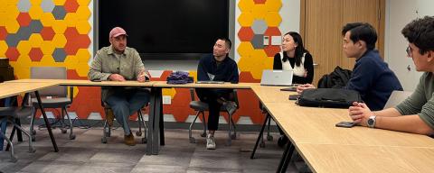 A speaker addresses a group of students seated around a U-shaped table in a modern classroom with a colorful honeycomb-patterned wall.