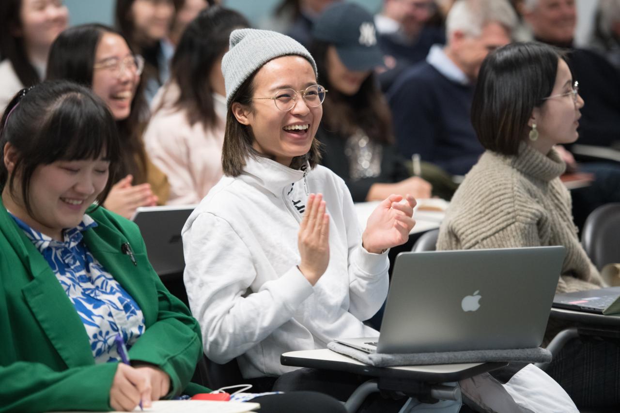 Students in a classroom smile and clap, with one wearing a beanie and glasses, seated at a desk with a laptop open.