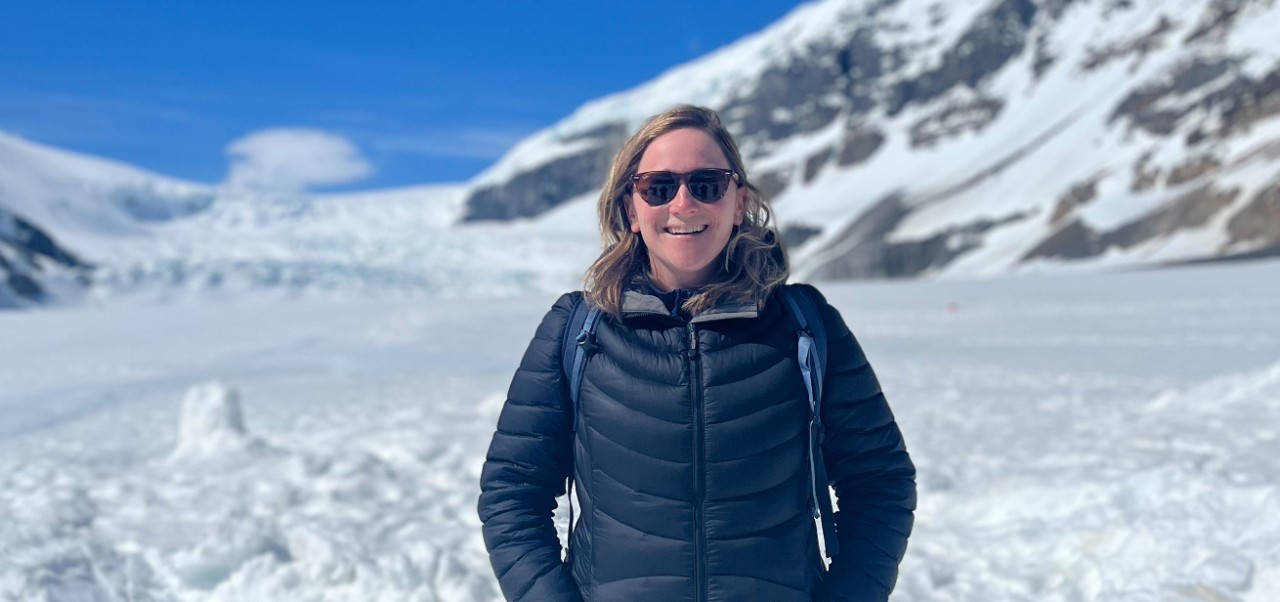 Caroline Kasper, NYU SPS Preston Robert Tisch Institute for Global Sport alumnus, smiles for the camera while standing in a snowy valley between mountains.