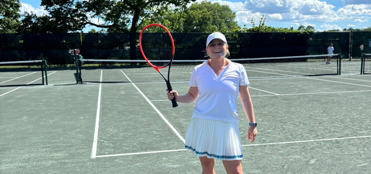 Caroline Kasper, NYU SPS Preston Robert Tisch Institute for Global Sport alumnus, takes a break from playing tennis to smile for the camera while holding her racket.