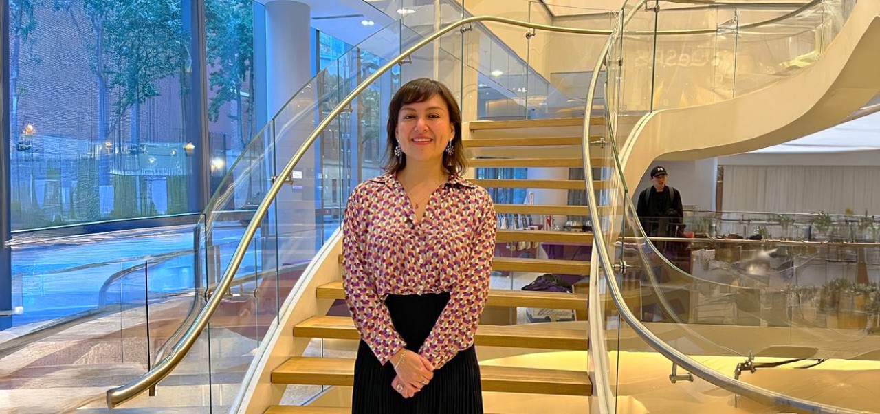 Gabriela Llerna, Division of Programs in Business at NYU SPS alumnus, smiles for the camera in front of a sleek spiral staircase in a window-lined atrium.